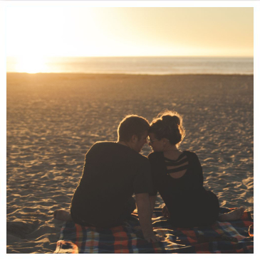 young couple sitting on a blanket on the beach sand, with the ocean and a setting sun behind them.